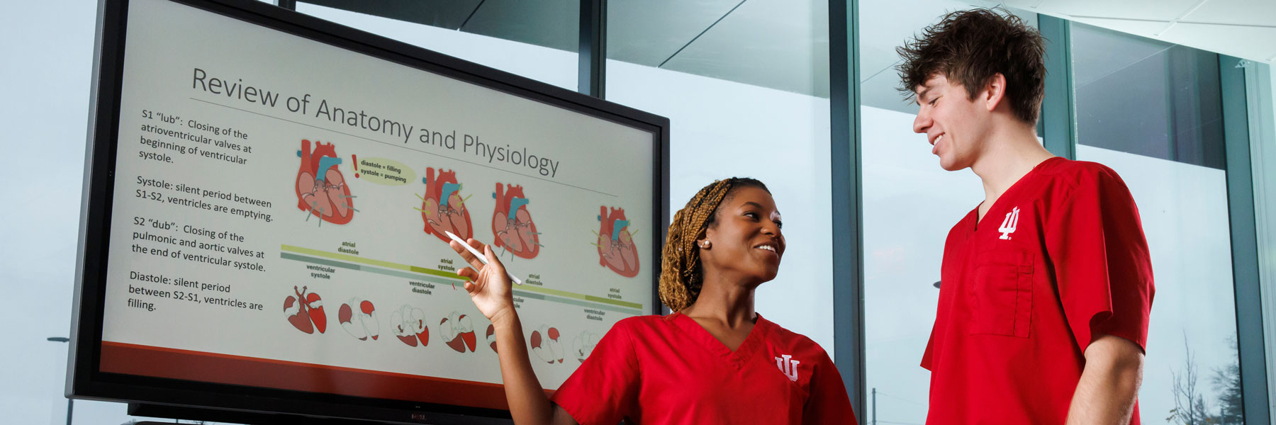 Two IU School of Nursing students discuss information on a television screen.