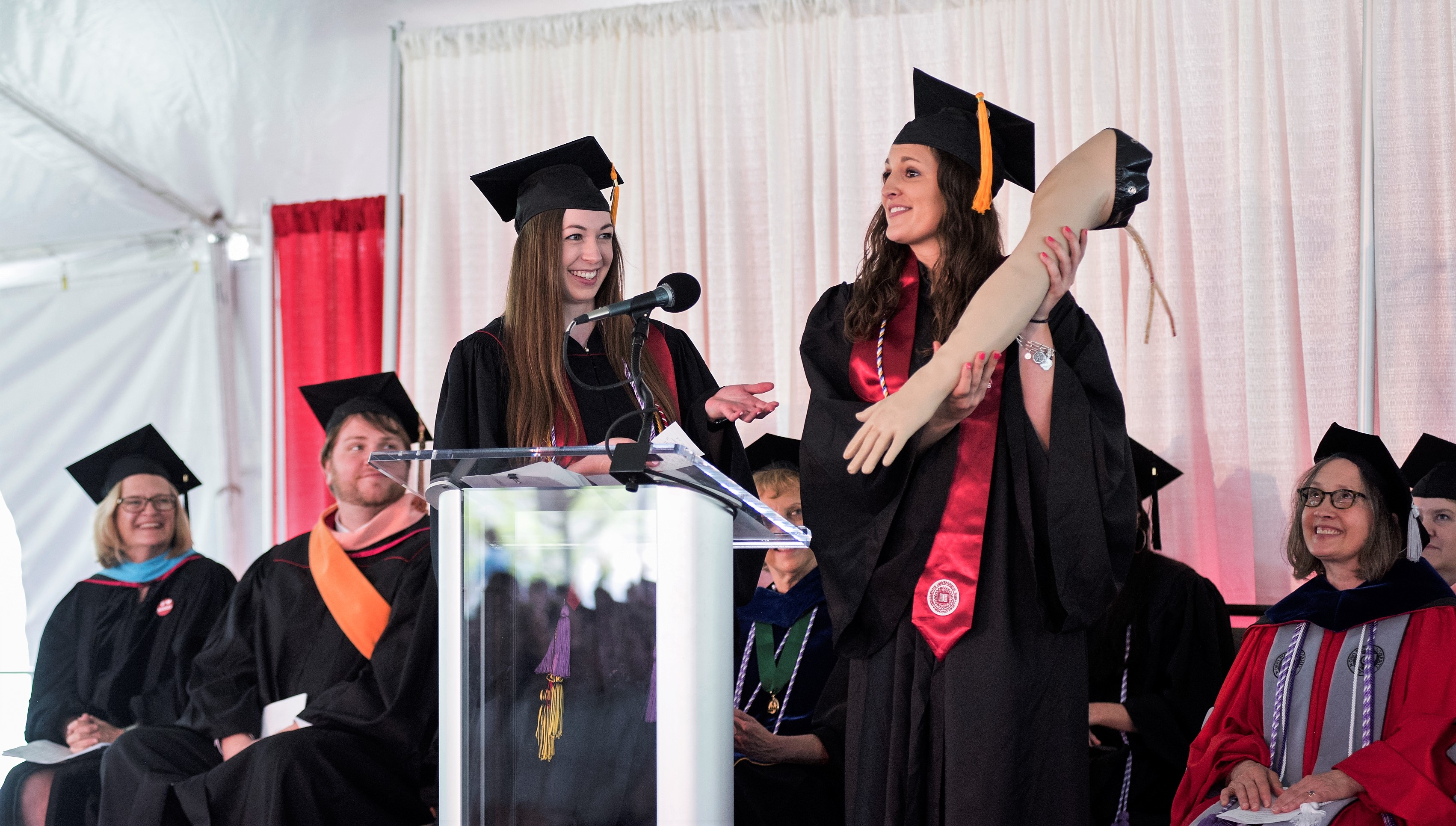 Two students in caps and gowns at graduation ceremony. One is holding a manikin's arm.