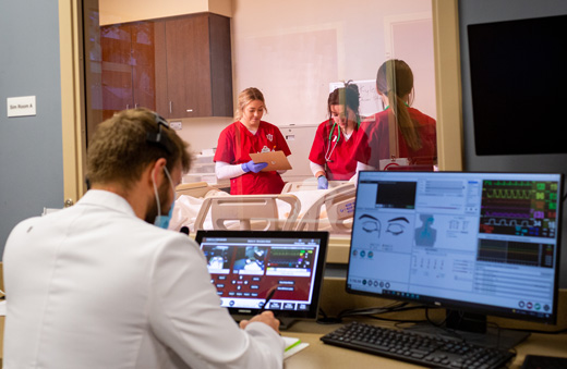 Three future IU nurses work in a simulation space while a person in a white lab coat observes